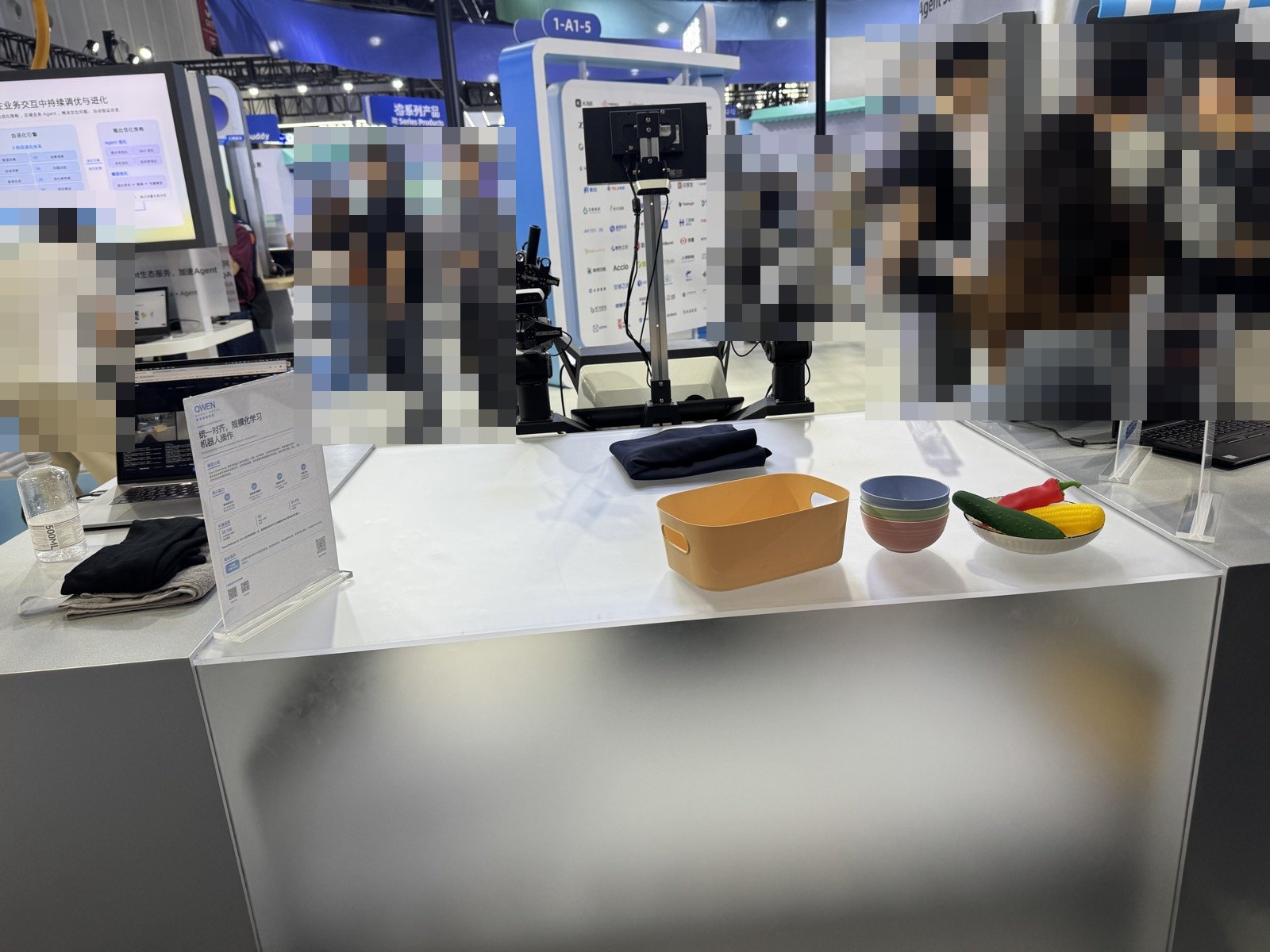 二本のロボットアームの前に、畳んだ布、オレンジのかご、重ねたお椀、野菜の模型が並ぶ展示台